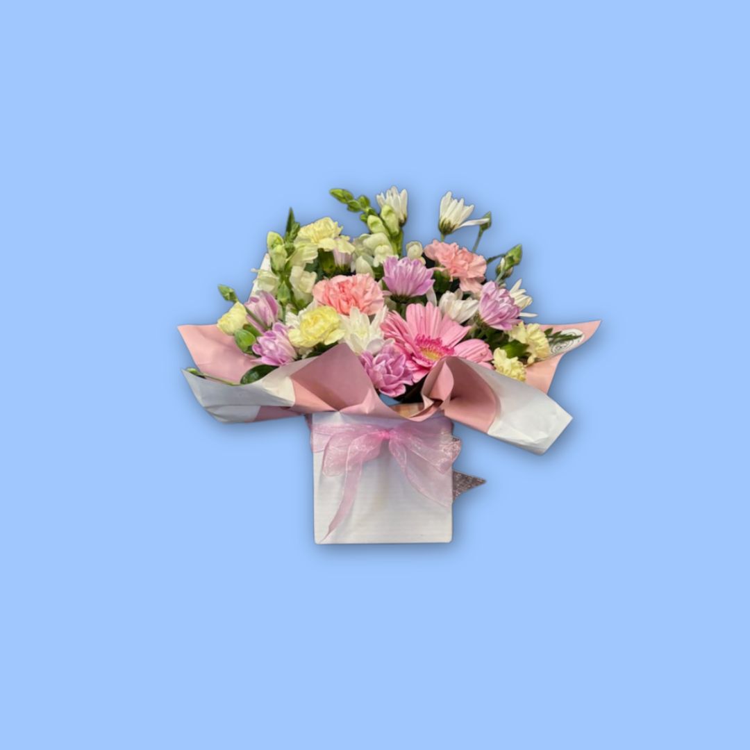 Small mixed pastels themed arrangement of flowers wrapped in pink paper displayed in a white posy box with a pink bow around it. picture has a sky blue background.