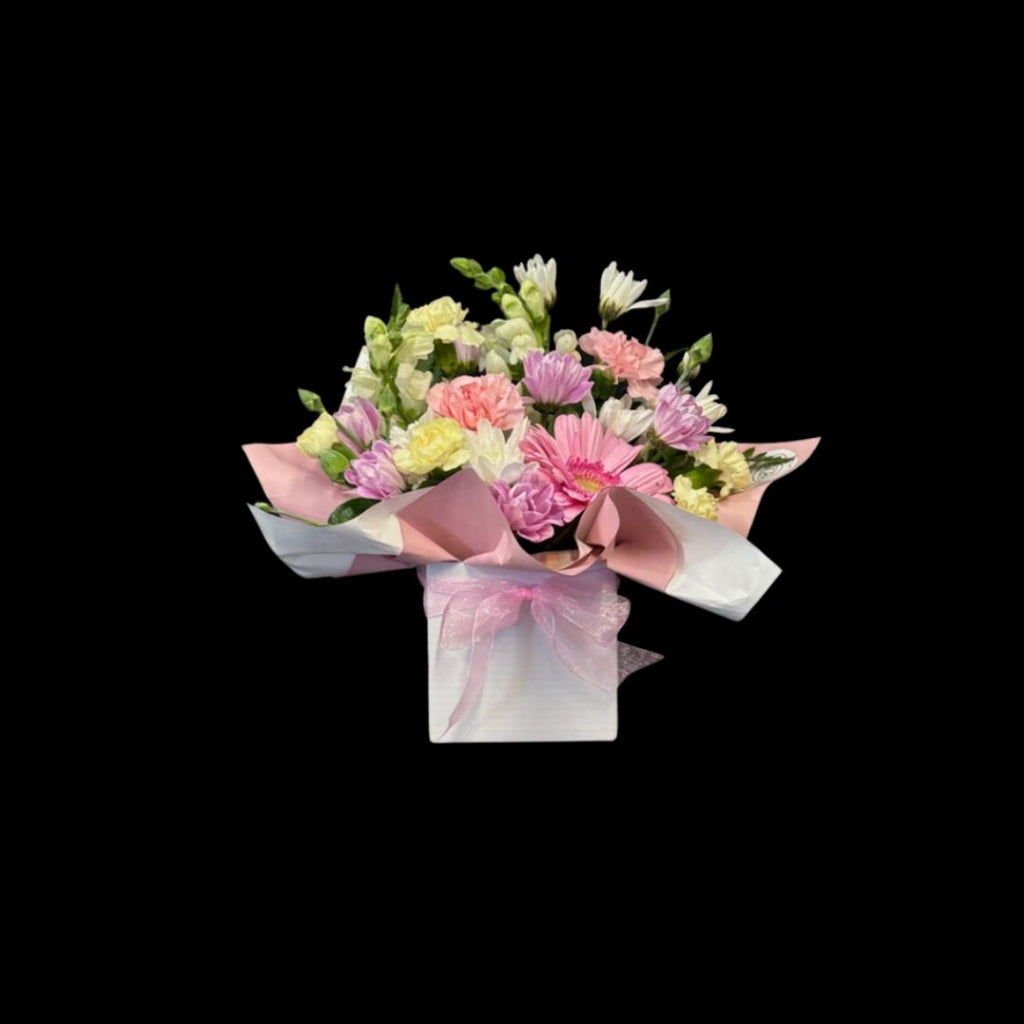 Small mixed pastels themed arrangement of flowers wrapped in pink paper displayed in a white posy box with a pink bow around it. picture has a black background.