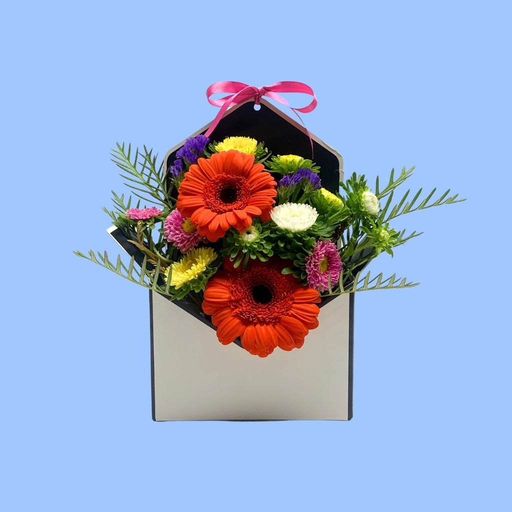 Front on view of Bright mixed Floral arrangement in an envelope shaped box with a pink bow 