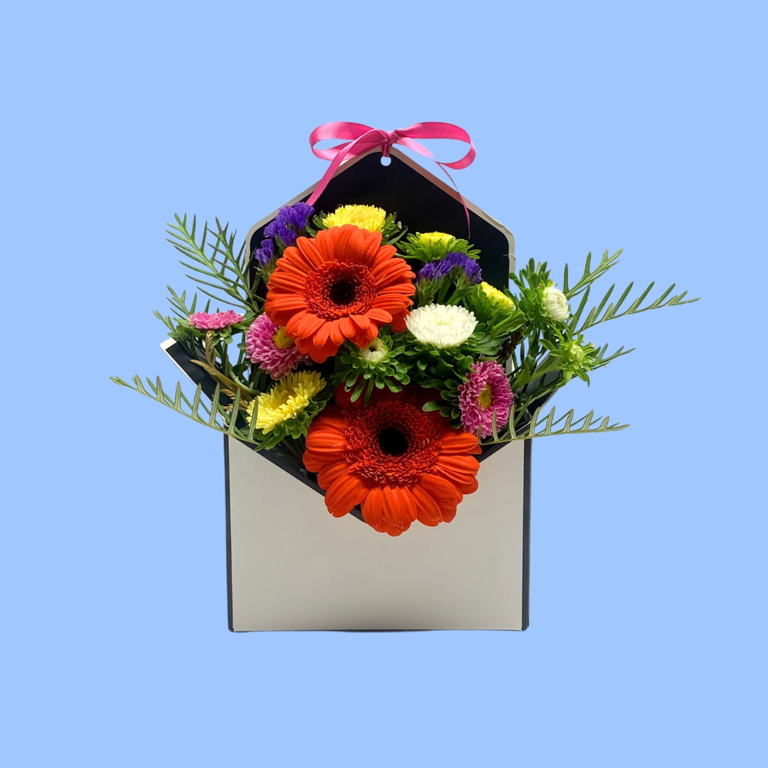 Front on view of Bright mixed Floral arrangement in an envelope shaped box with a pink bow 