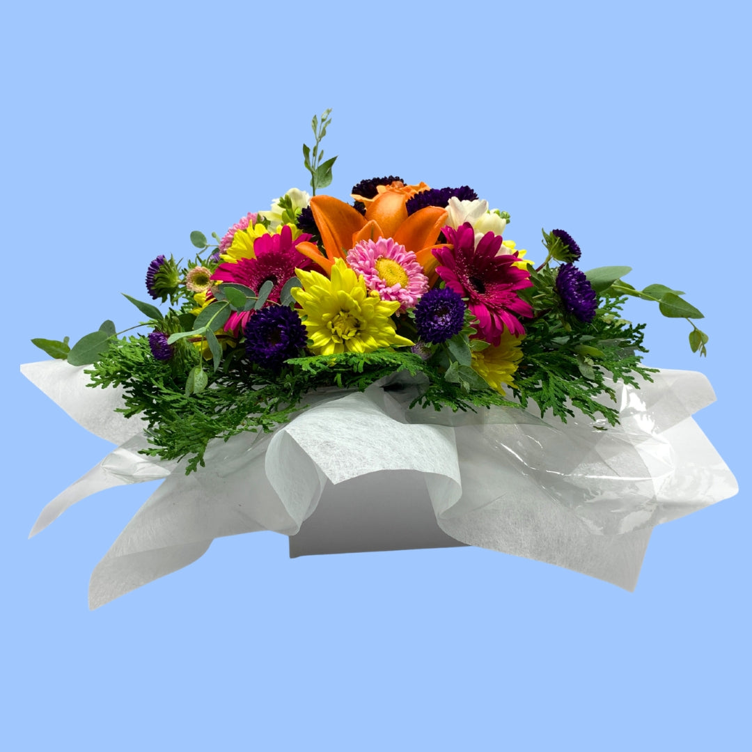 Side view of low arrangement of mixed bright flowers against greenery and white tissue in a white box