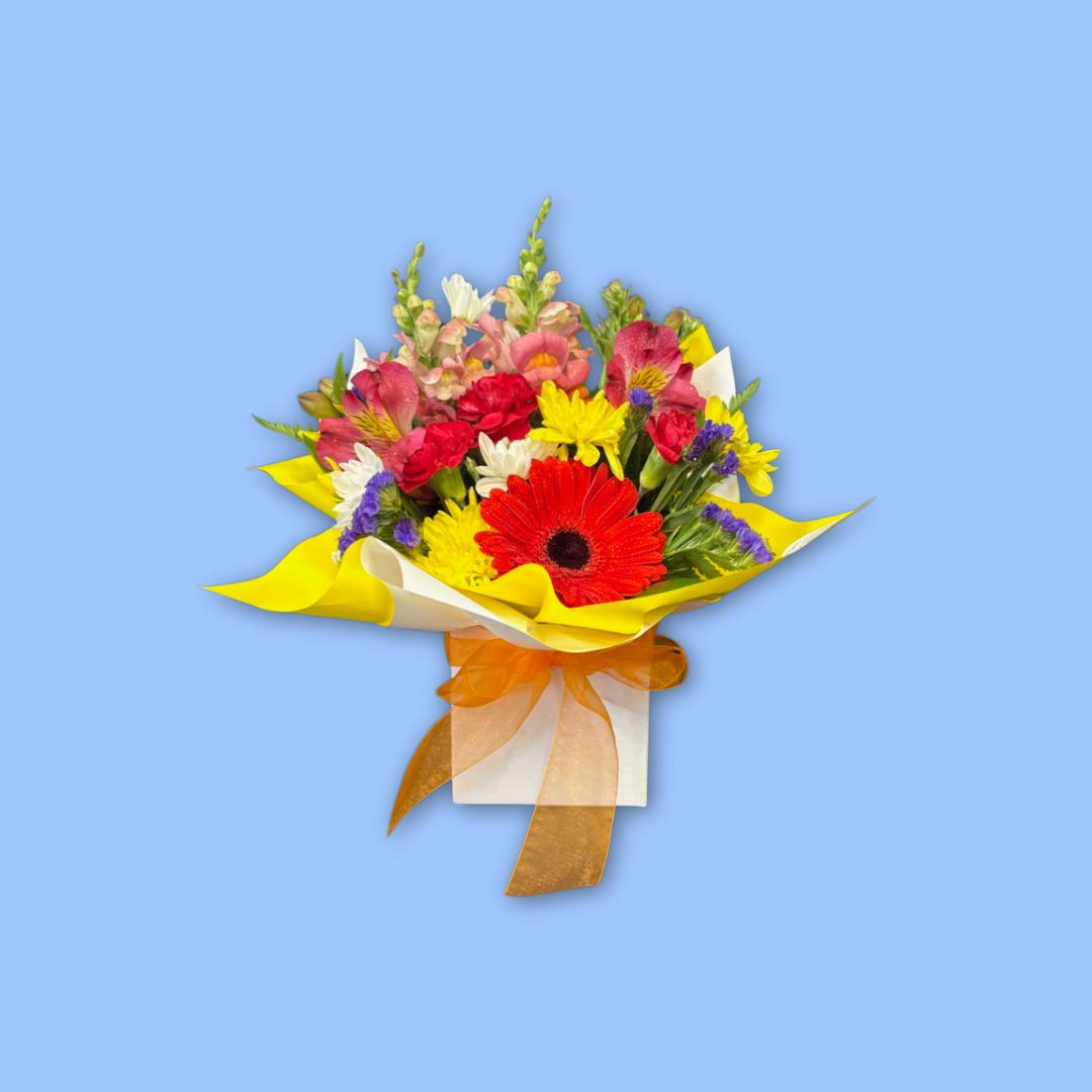 Small mixed bright themed arrangement of flowers in yellow paper displayed in a white posy box with an orange bow around it.