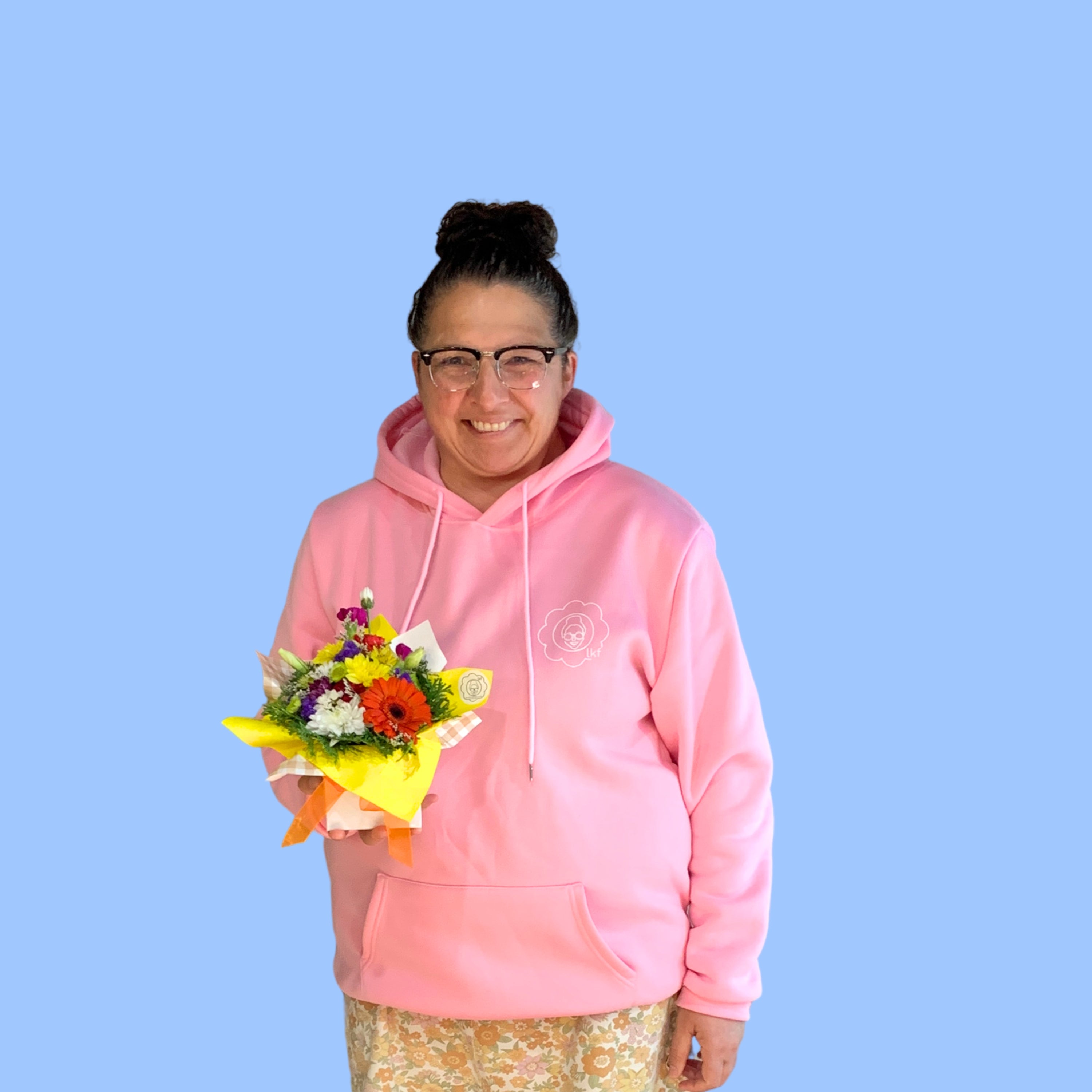 Laura King holding a Small mixed bright themed arrangement of flowers in yellow paper displayed in a white posy box with an orange bow around it.