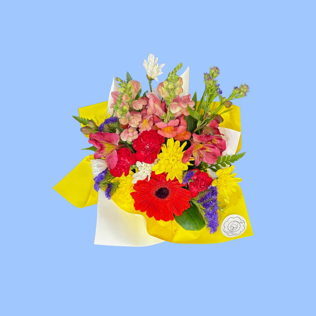 A top view of Small mixed bright themed arrangement of flowers in yellow paper displayed in a white posy box with an orange bow around it.