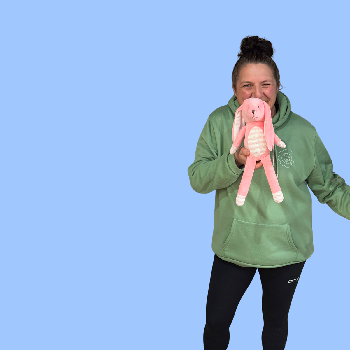 A woman wearing a sage green hoodie and black leggings holds a pink plush bunny against her face, set against a solid light blue background.