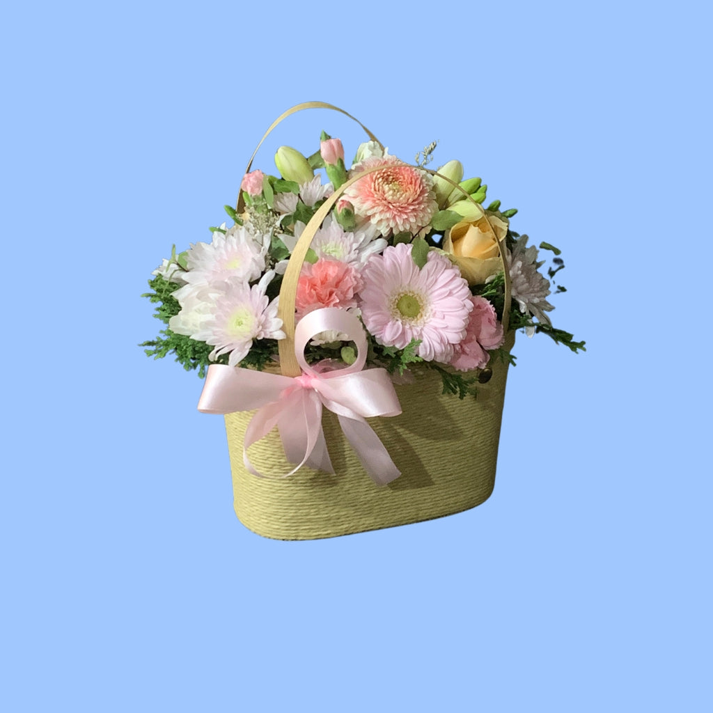Arrangement of pastels coloured flowers in small woven style basket with handles and a pink bow