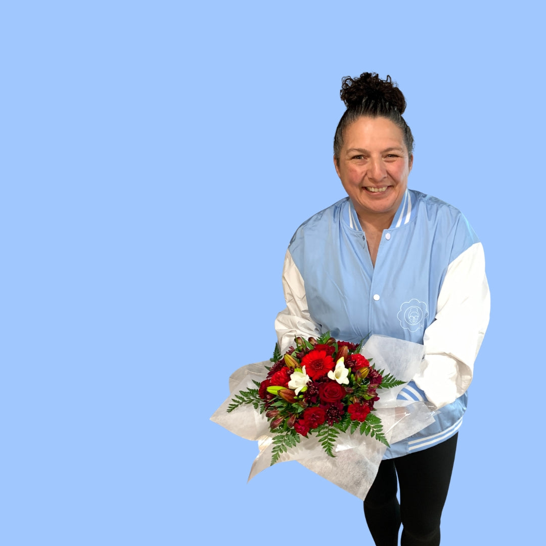 Laura King holding a floral box arrangement with red roses, burgundy gerbera daisies, white lilies, and green ferns, wrapped in white tissue paper.