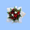 Top view of a floral box arrangement with red roses, burgundy gerbera daisies, white lilies, and green ferns, wrapped in white tissue paper.