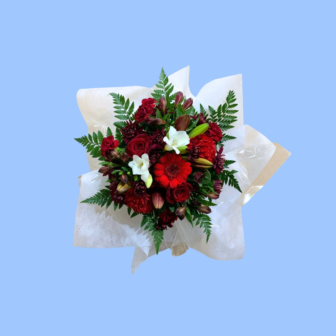 Top view of a floral box arrangement with red roses, burgundy gerbera daisies, white lilies, and green ferns, wrapped in white tissue paper.