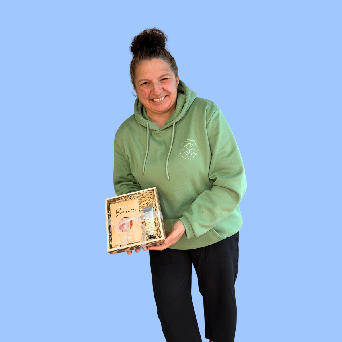 Laura King florist holding a gift box set containing a tube of charlotte's lab Hand Cream and a pack of Rosé gummy bears bears, presented in a wooden box with a white ribbon around it on a solid blue background. 