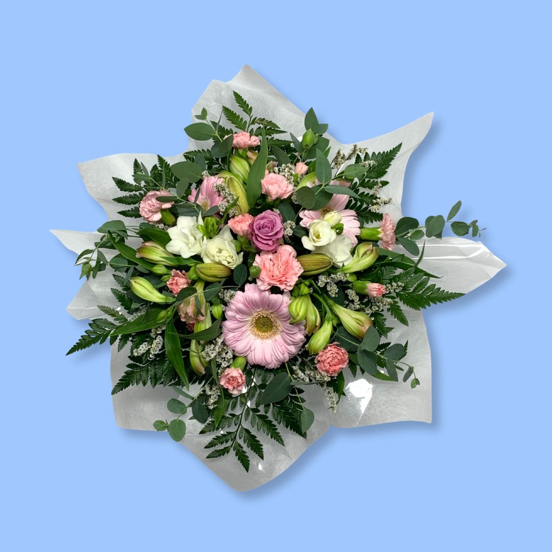 Top down view of a low arrangement of mixed softer pastel coloured flowers against greenery and wrapped in tissue.