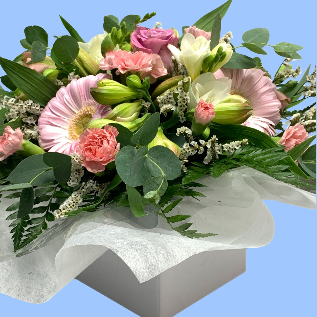side view view of a low arrangement of mixed softer pastel coloured flowers against greenery and wrapped in tissue and in a white box