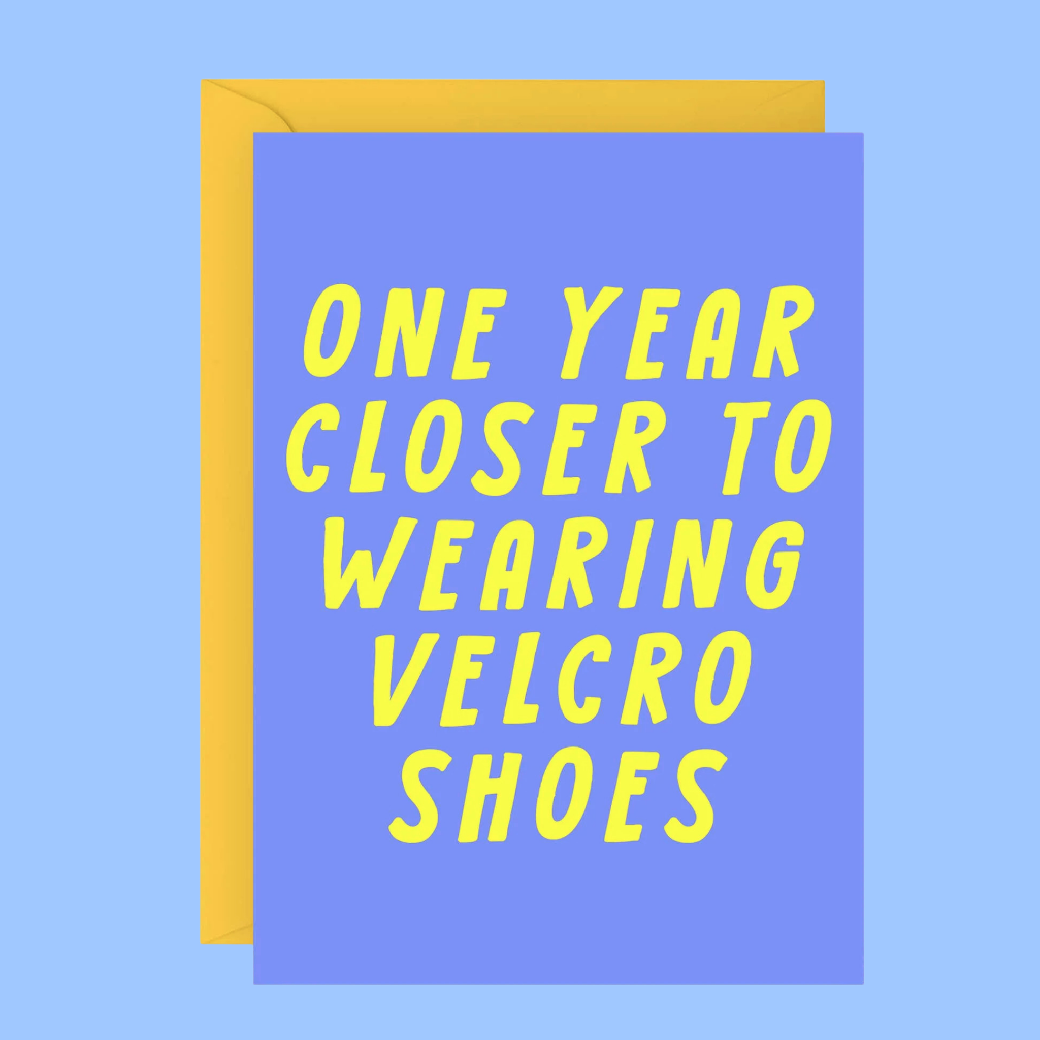 A square greeting card with a lavender-blue background and bold yellow text reading 'ONE YEAR CLOSER TO WEARING VELCRO SHOES,' shown with a yellow envelope behind it on a light blue background.